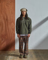 Full-length front view of model wearing Uskees 'vine green' coloured buttoned smock, paired with brown check trousers and bear hunter-style hat.