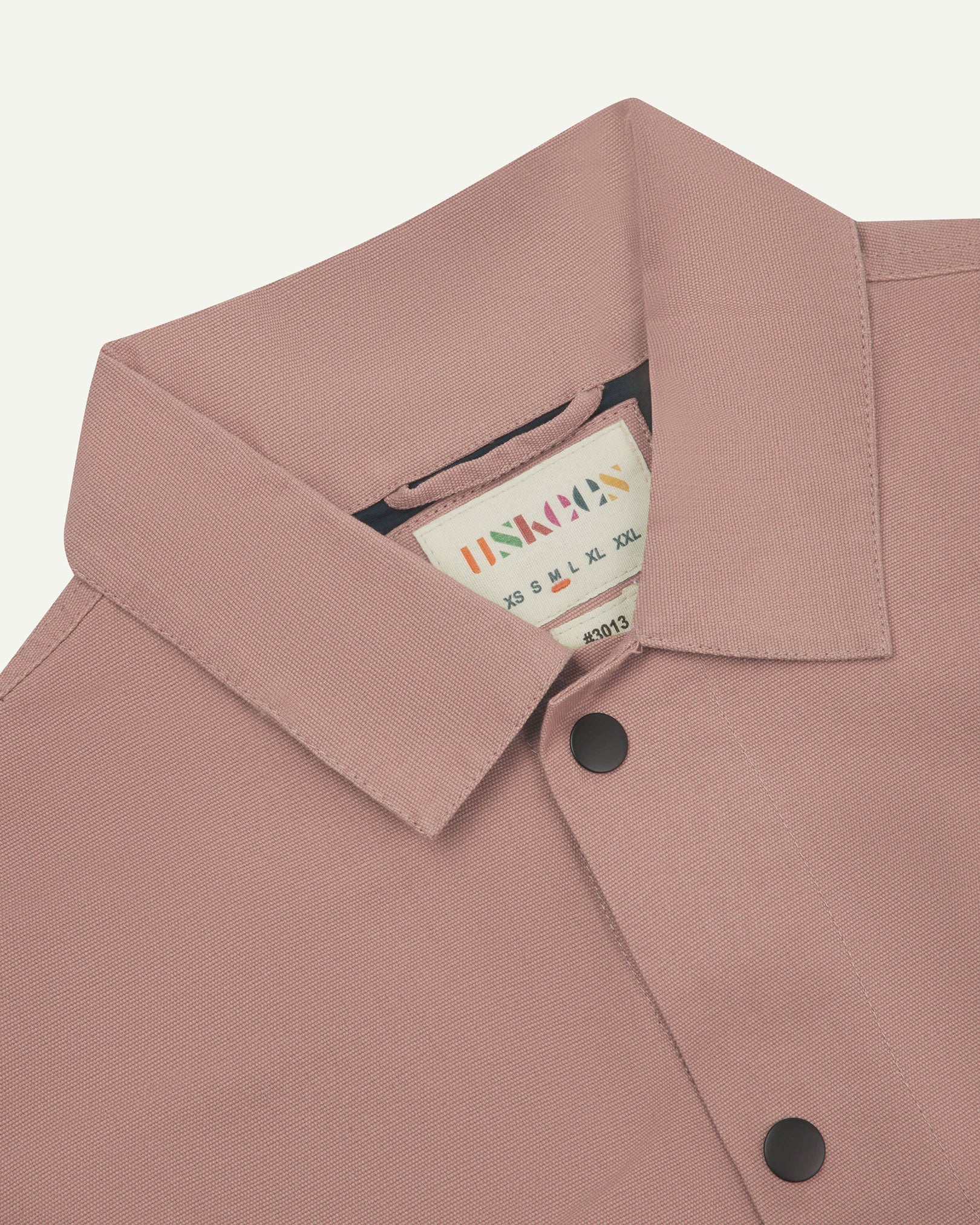 Neck view of Uskees 3013 dusty pink organic cotton coach jacket with focus on collar, contrast inner yoke, Uskees brand label and popper buttons.