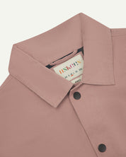 Neck view of Uskees 3013 dusty pink organic cotton coach jacket with focus on collar, contrast inner yoke, Uskees brand label and popper buttons.