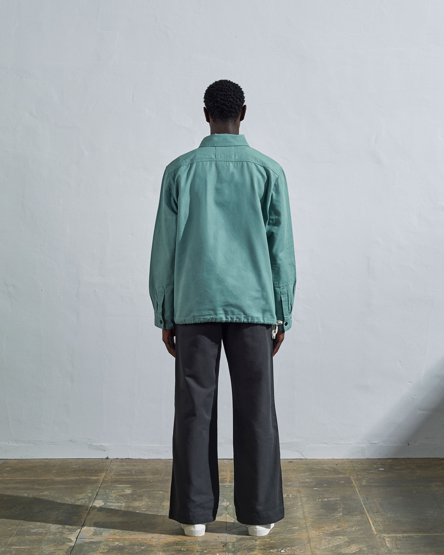 Full-length back view of model wearing Uskees Coach's jacket in eucalyptus blue-green, illustrating reinforced elbow and simple, boxy silhouette. 