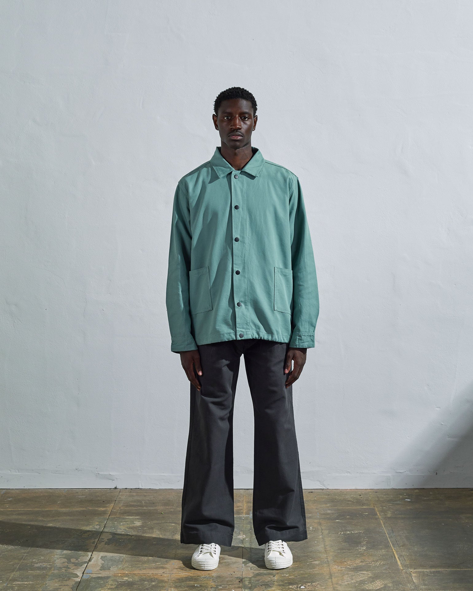 Full-length front view of model wearing Uskees Coach's jacket in eucalyptus blue-green. Paired with dark coloured pants.