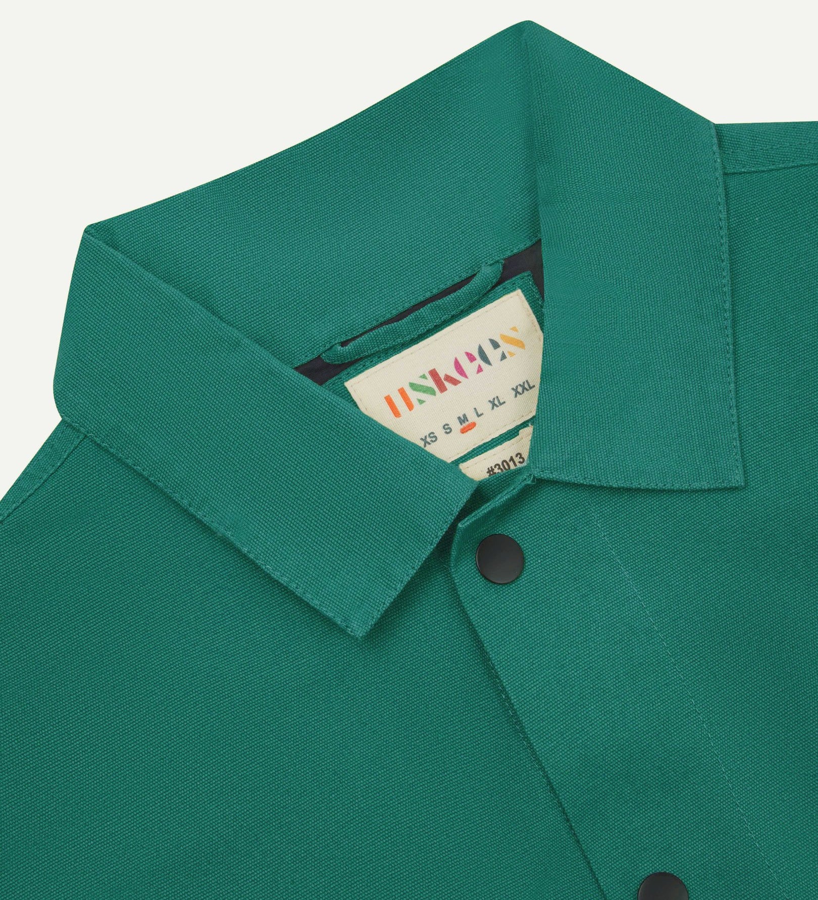 Neck view of Uskees 3013 foam green organic cotton coach jacket with focus on collar, contrast inner yoke, Uskees brand label and popper buttons.