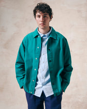 Male model wearing Uskees foam green organic cotton coach jacket undone over a pale blue shirt.