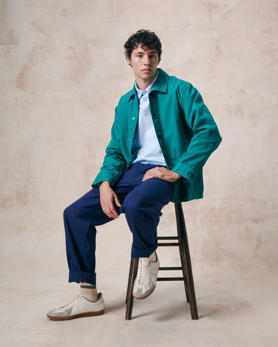 Model seated on a stool wearing Uskees foam green organic cotton coach jacket undone with dark blue trousers