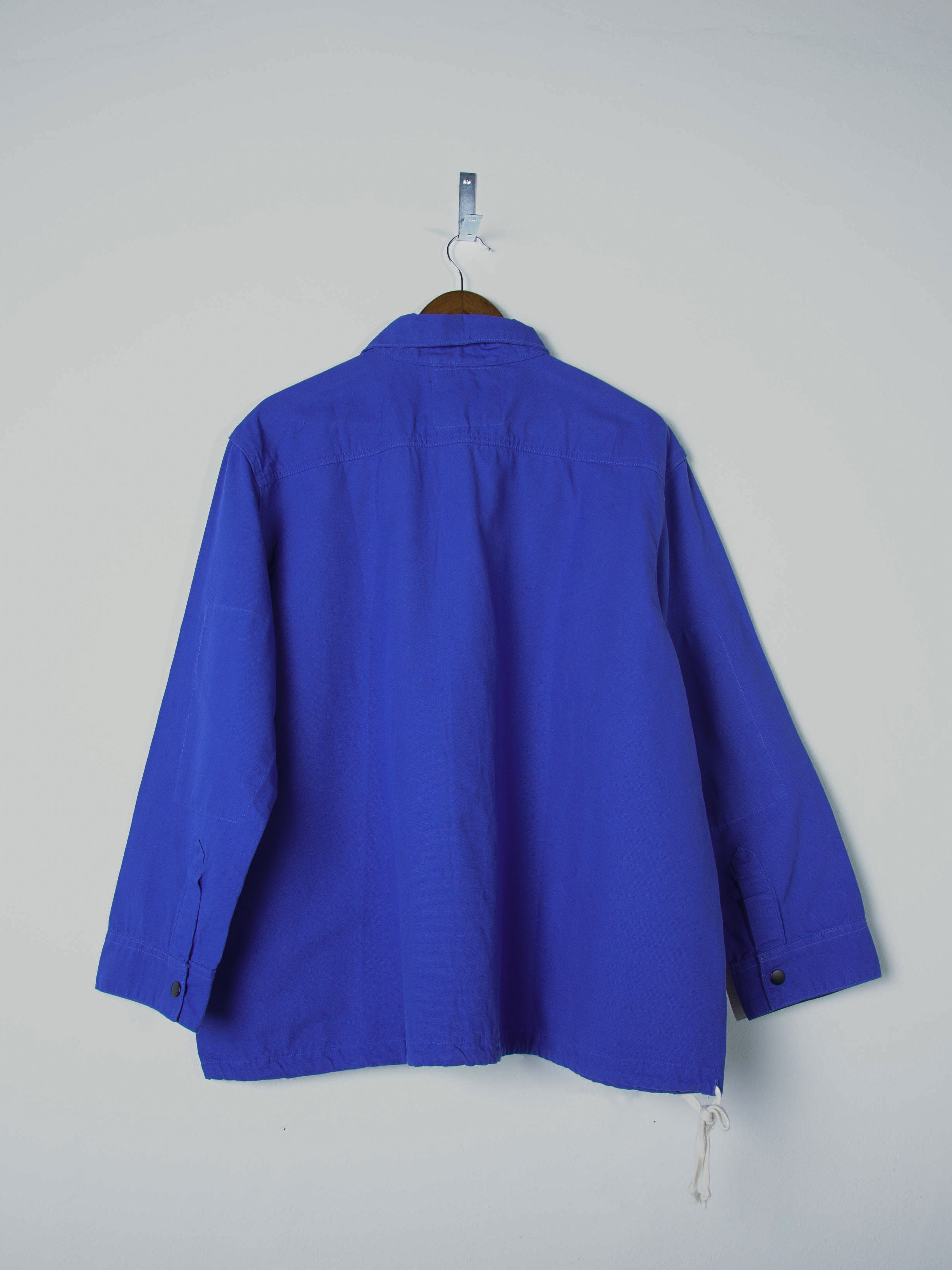 Reverse view of Uskees ultra blue organic cotton coach jacket, illustrating reinforced elbows, drawstring base and simple, boxy silhouette. 