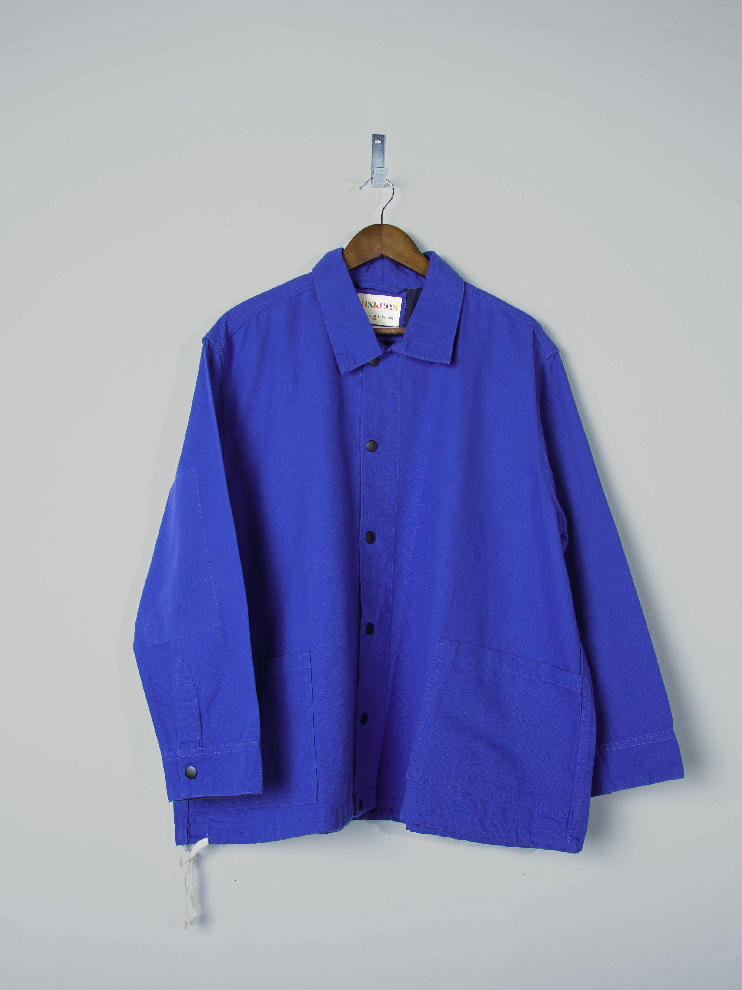 Front view of Uskees ultra blue organic cotton coach jacket with 2 patch pockets, popper fastening and contrast yoke. Presented on hanger.