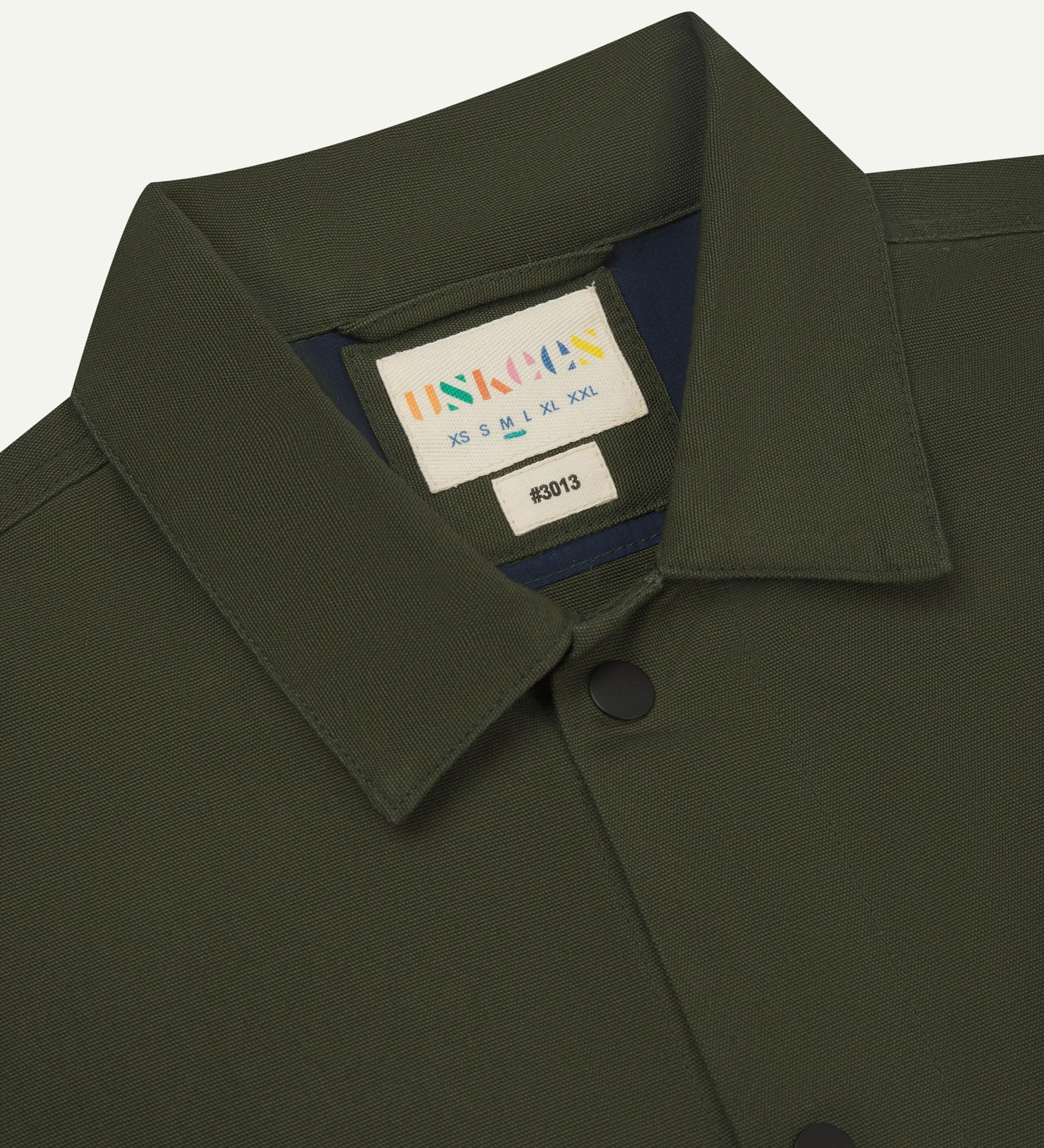 Neck view of Uskees 3013 vine green organic cotton coach jacket with focus on collar, contrast inner yoke, Uskees brand label and popper buttons.