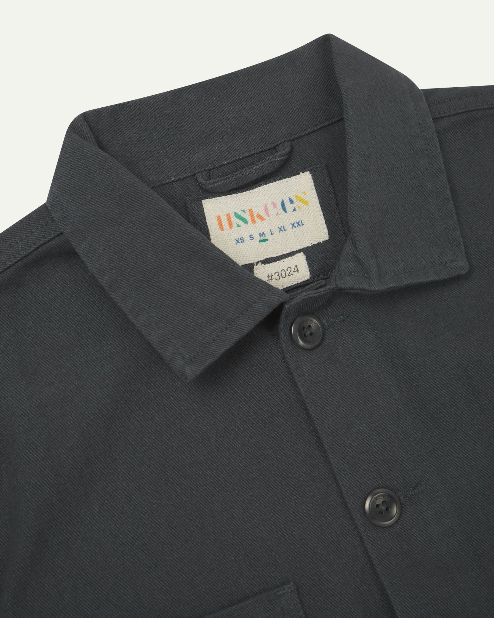 Close-up top-half view of #3024, charcoal-grey organic cotton drill over shirt. With focus on collar, Uskees brand label and corozo buttons.