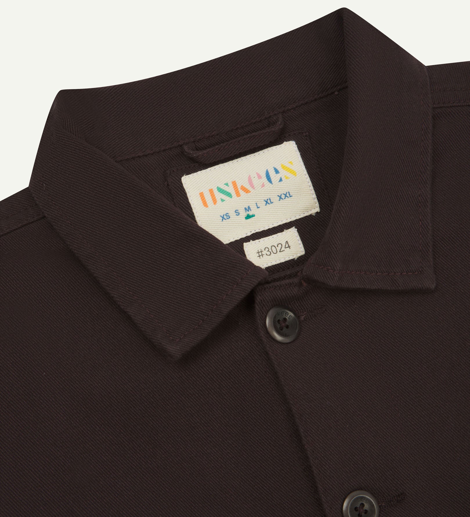 Close-up top-half view of #3024, burgundy-brown-coloured organic cotton-drill over shirt. With focus on collar, Uskees brand label and corozo buttons.
