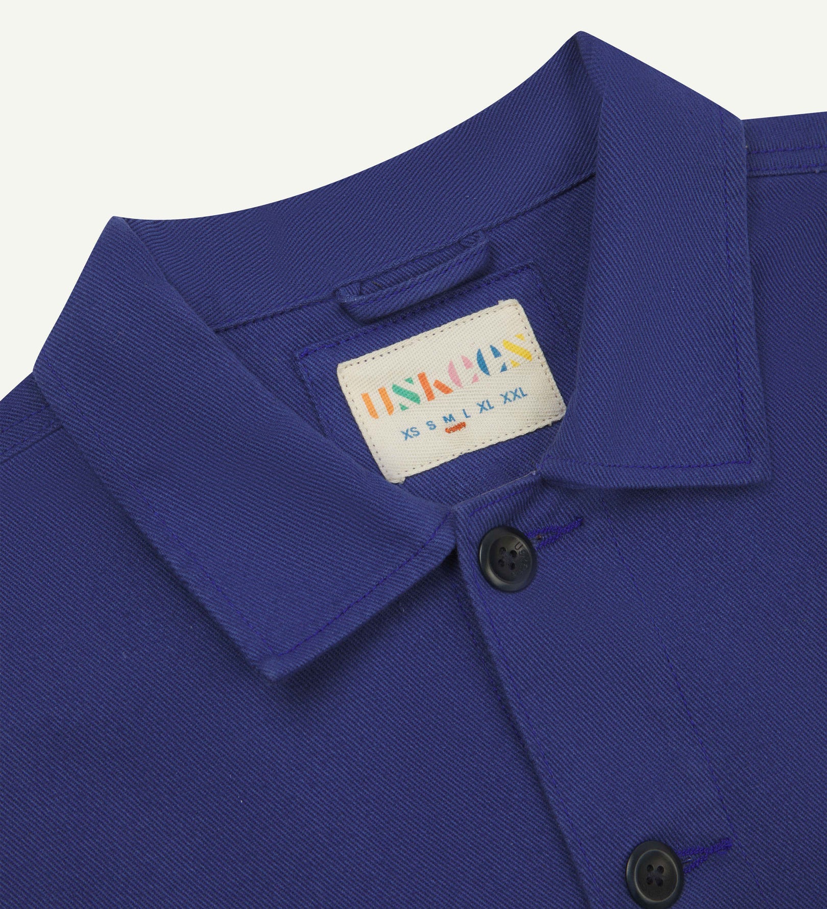Close-up top-half view of #3024, ultra blue-coloured organic cotton-drill over shirt. With focus on collar, Uskees brand label and corozo buttons.