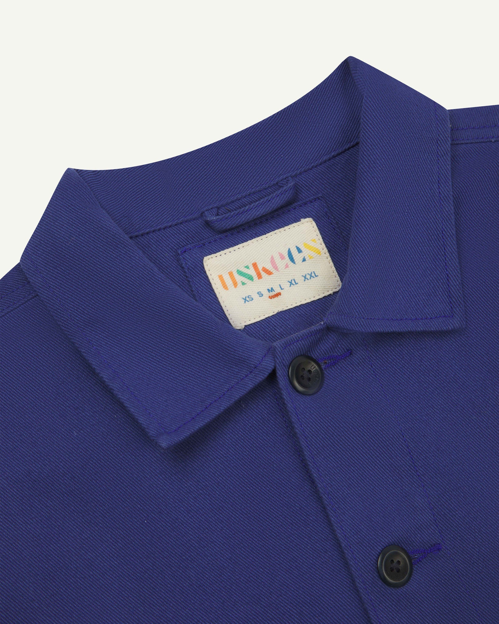 Close-up top-half view of #3024, ultra blue-coloured organic cotton-drill over shirt. With focus on collar, Uskees brand label and corozo buttons.