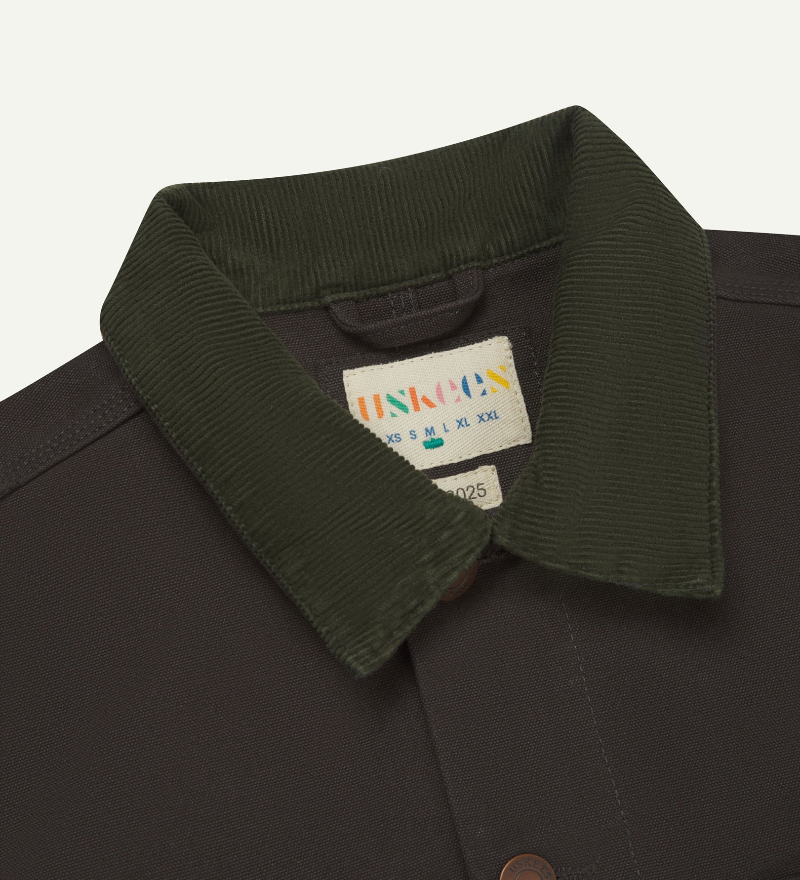 Front close-up shot of Uskees cotton canvas chore jacket for men in charcoal-grey. Clear view of the green corduroy collar, brand label and buttons