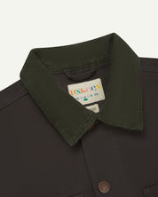 Front close-up shot of Uskees cotton canvas chore jacket for men in charcoal-grey. Clear view of the green corduroy collar, brand label and buttons