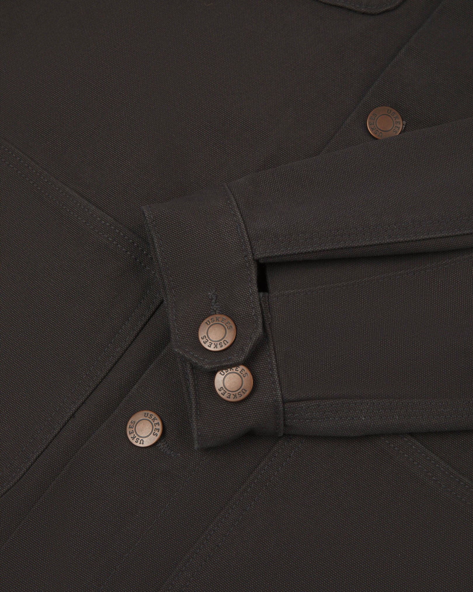 Detail shot of Uskees men's canvas chore jacket in charcoal-grey with a green collar showing the adjustable cuff, sleeve and pockets in close-up 