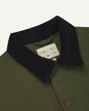 Close-up view of Uskees cotton canvas chore jacket for men in coriander green showing the dark blue cord collar, brand label and metal buttons 