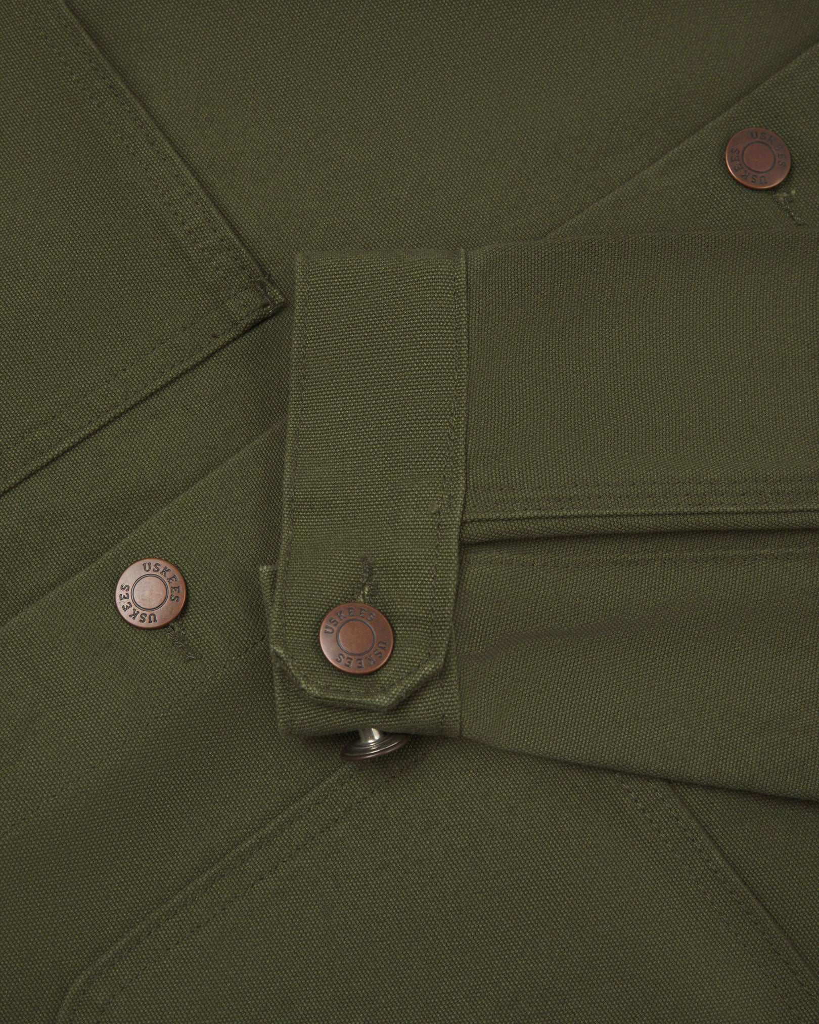 Close-up view of Uskees cotton canvas chore jacket in coriander green showing the adjuster buttons on the cuffs