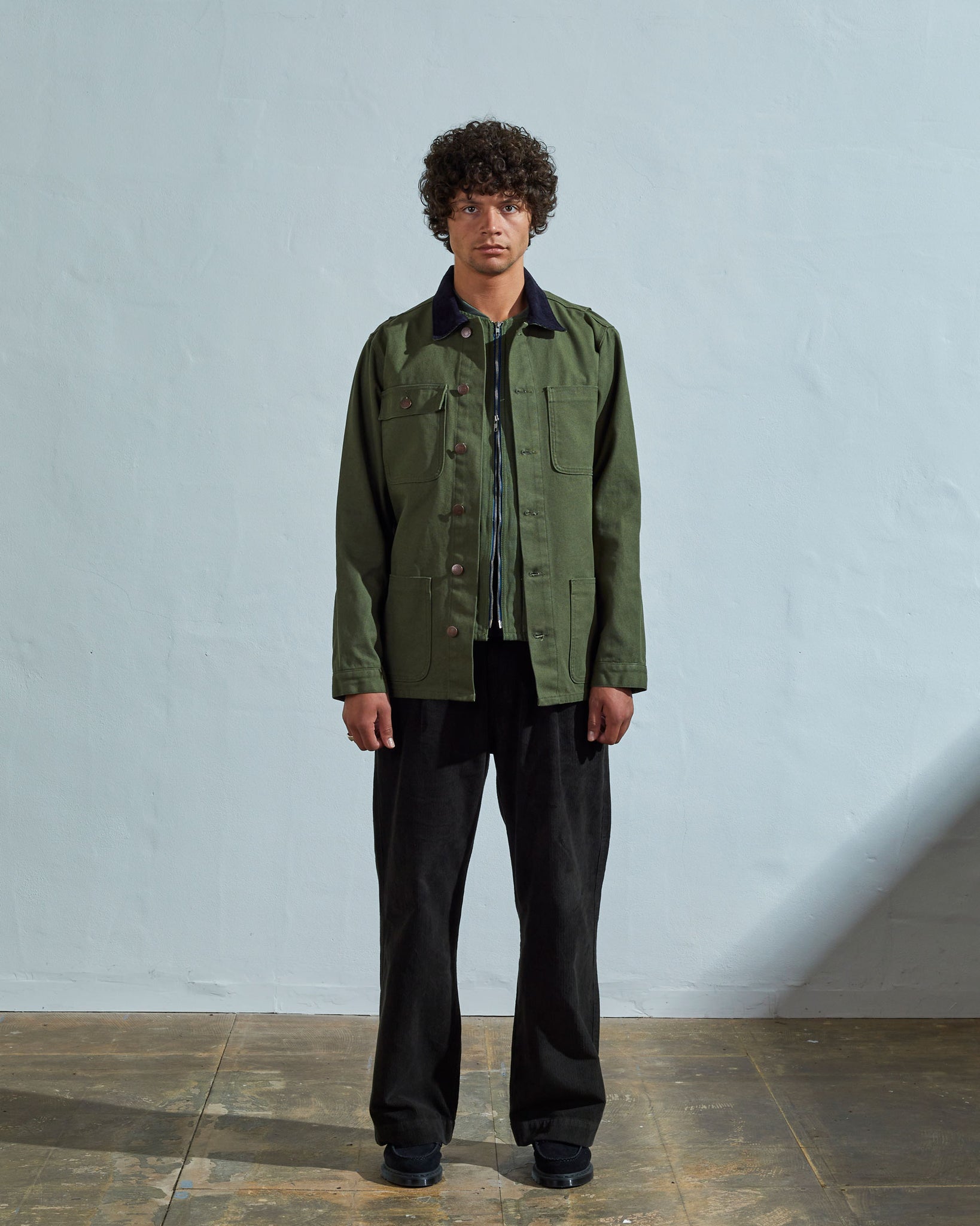 Full-length front view of model wearing unbuttoned #3025 coriander-green organic canvas chore jacket paired with black Uskees pants.