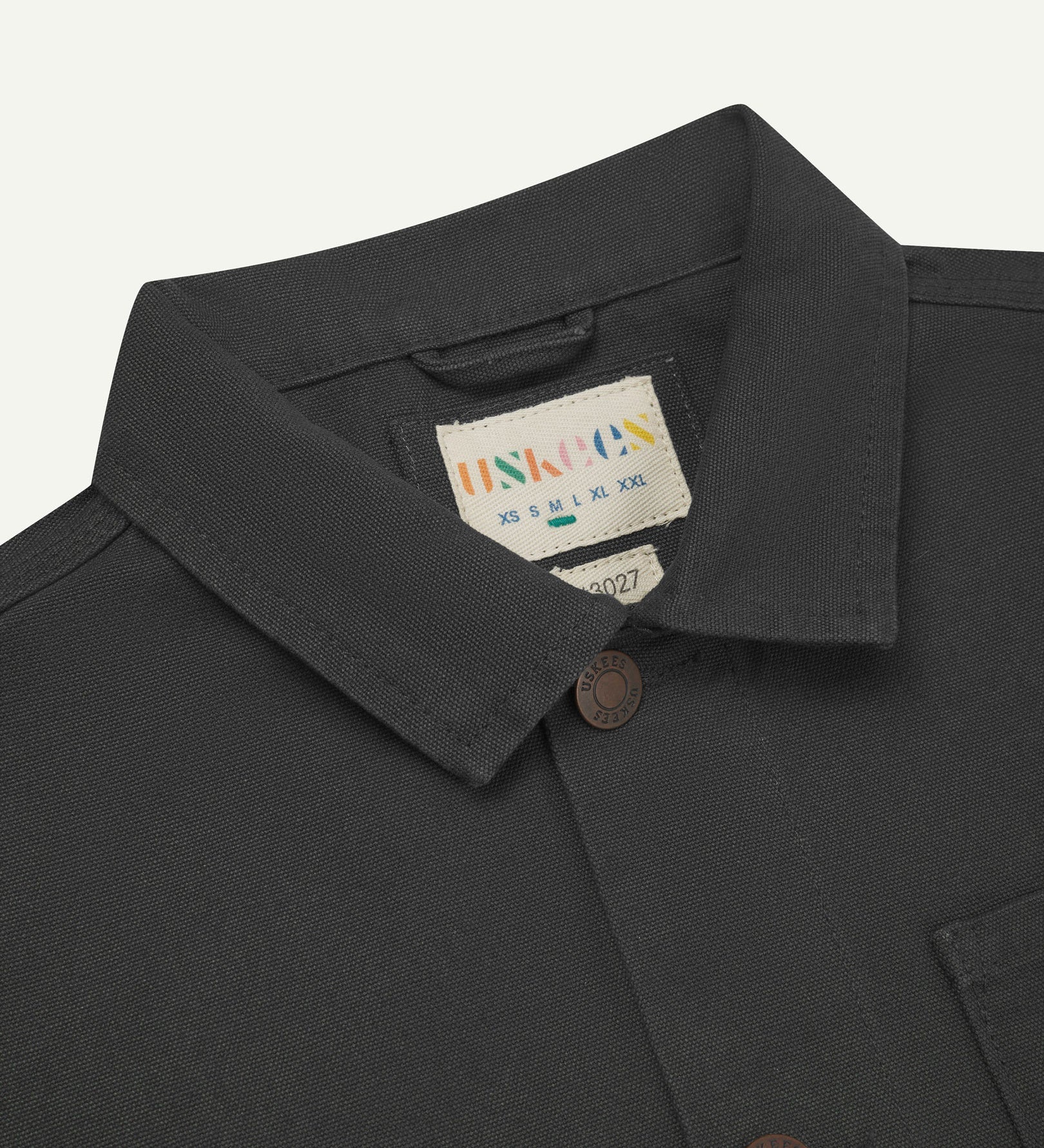 Front close-up view of Uskees dark grey canvas men's overshirt presented buttoned up showing the metal buttons, collar and brand label.