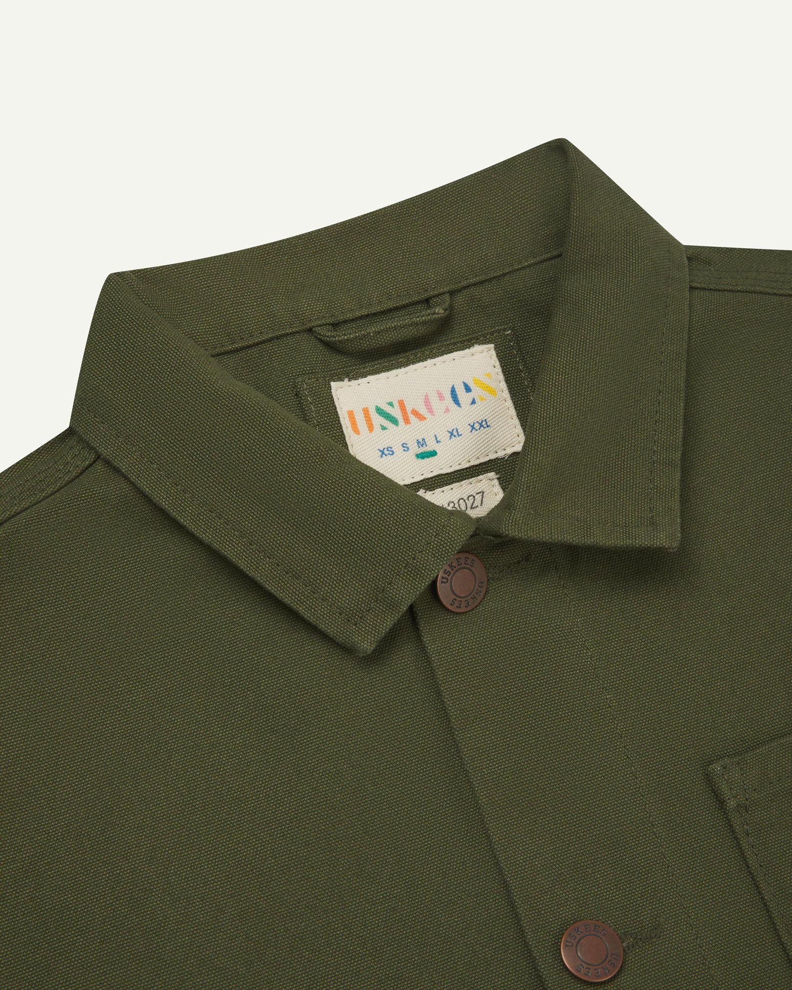 Front close-up view of Uskees coriander-green canvas men's overshirt presented buttoned up showing the metal buttons, collar and brand label.