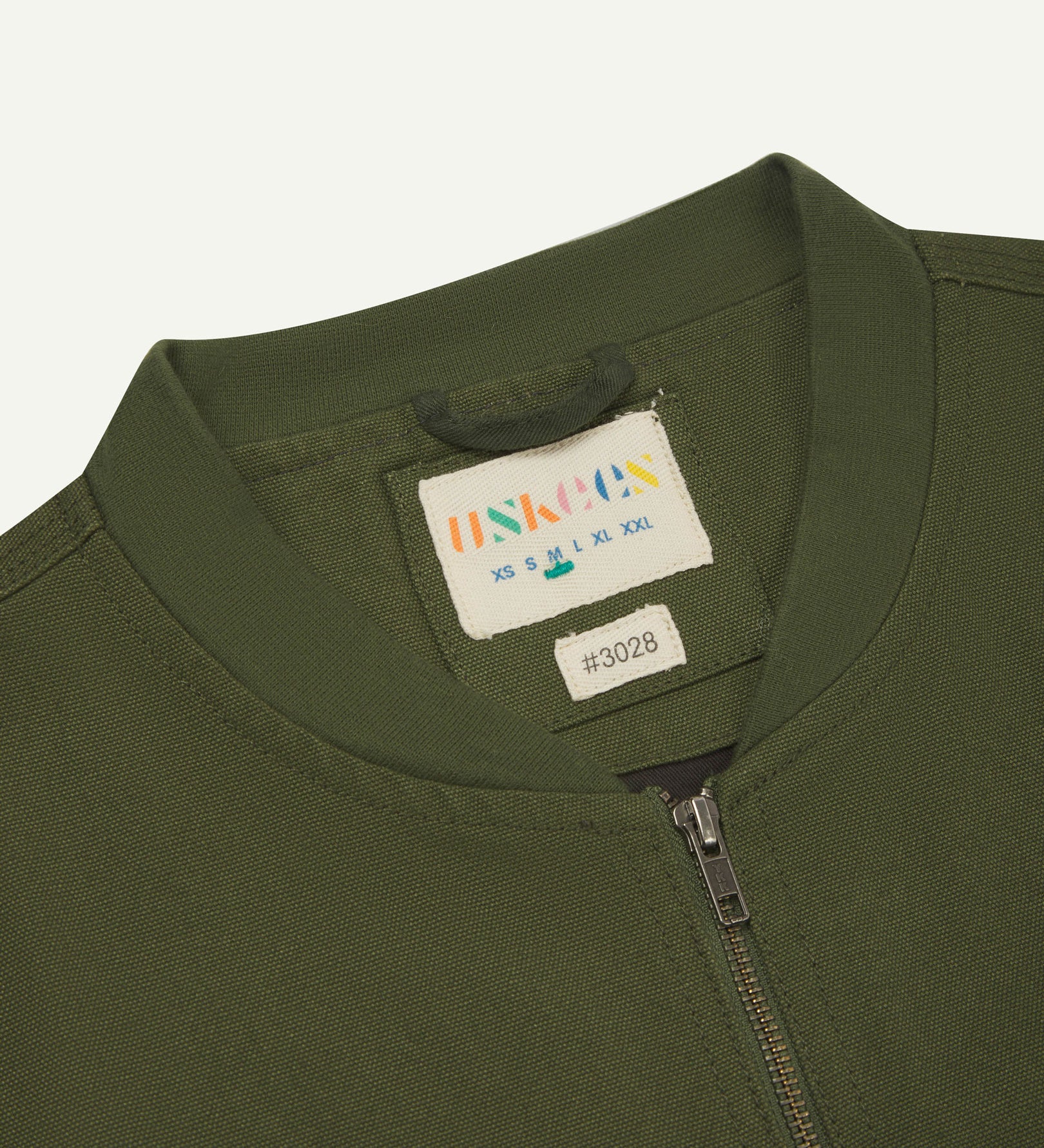 Close-up flat shot of Uskees coriander-green gilet-type zip front waistcoat showing jersey cotton collar neck, zip and branding label.