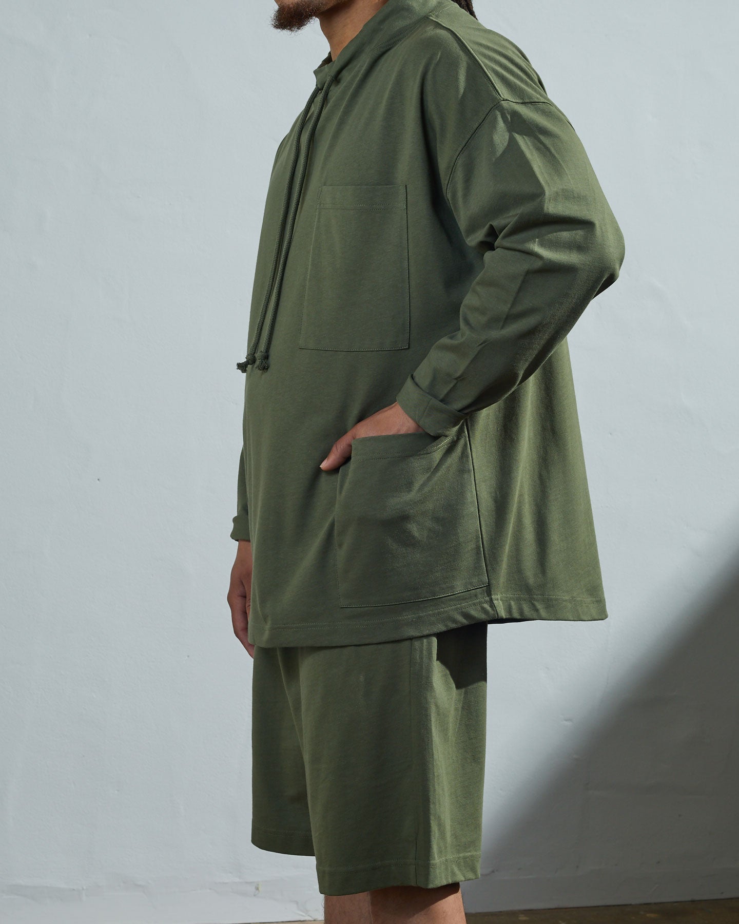 Side-view of model wearing army green organic cotton #3032 jersey tie neck smock by Uskees with hand in large hip patch pocket. Paired with matching #7007 shorts.