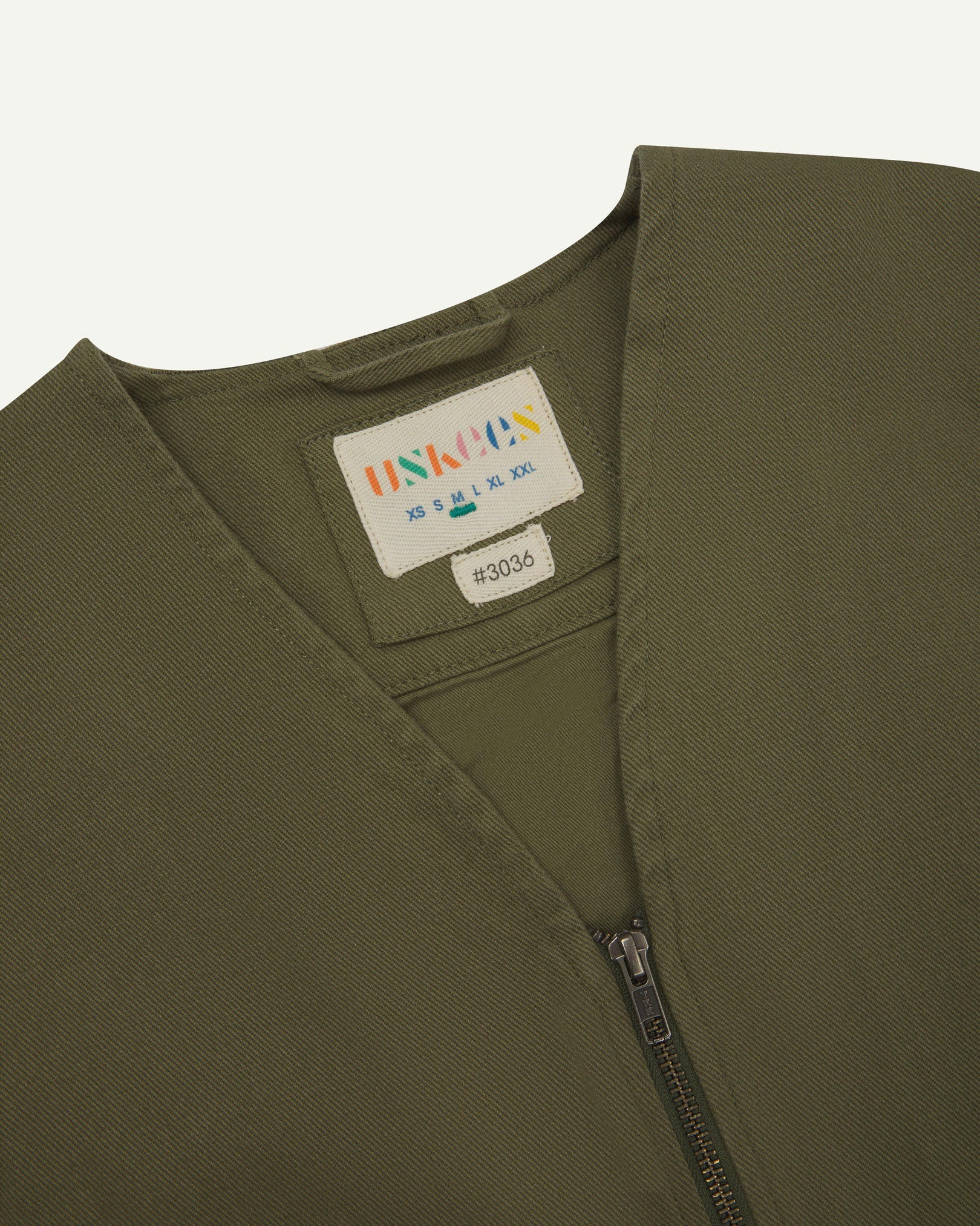 Close-up top-half view of #3036 moss-green organic cotton-drill vest. With focus on v-neck, contrasting black lining and Uskees branding label.