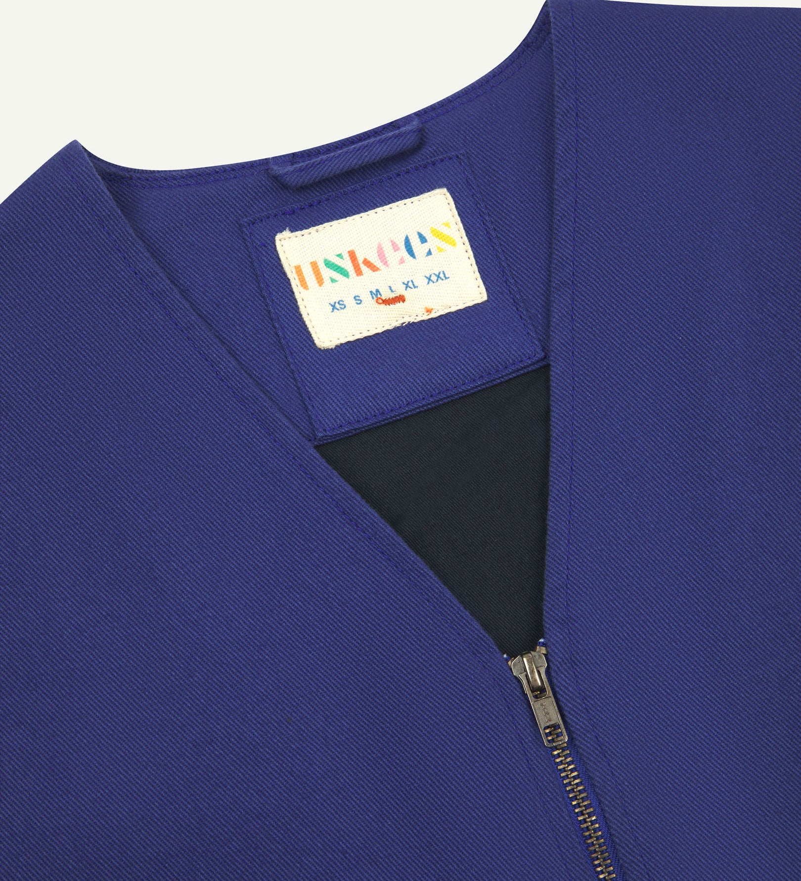 Close-up top-half view of #3036 ultra blue organic cotton-drill vest. With focus on v-neck, contrasting black lining and Uskees branding label.