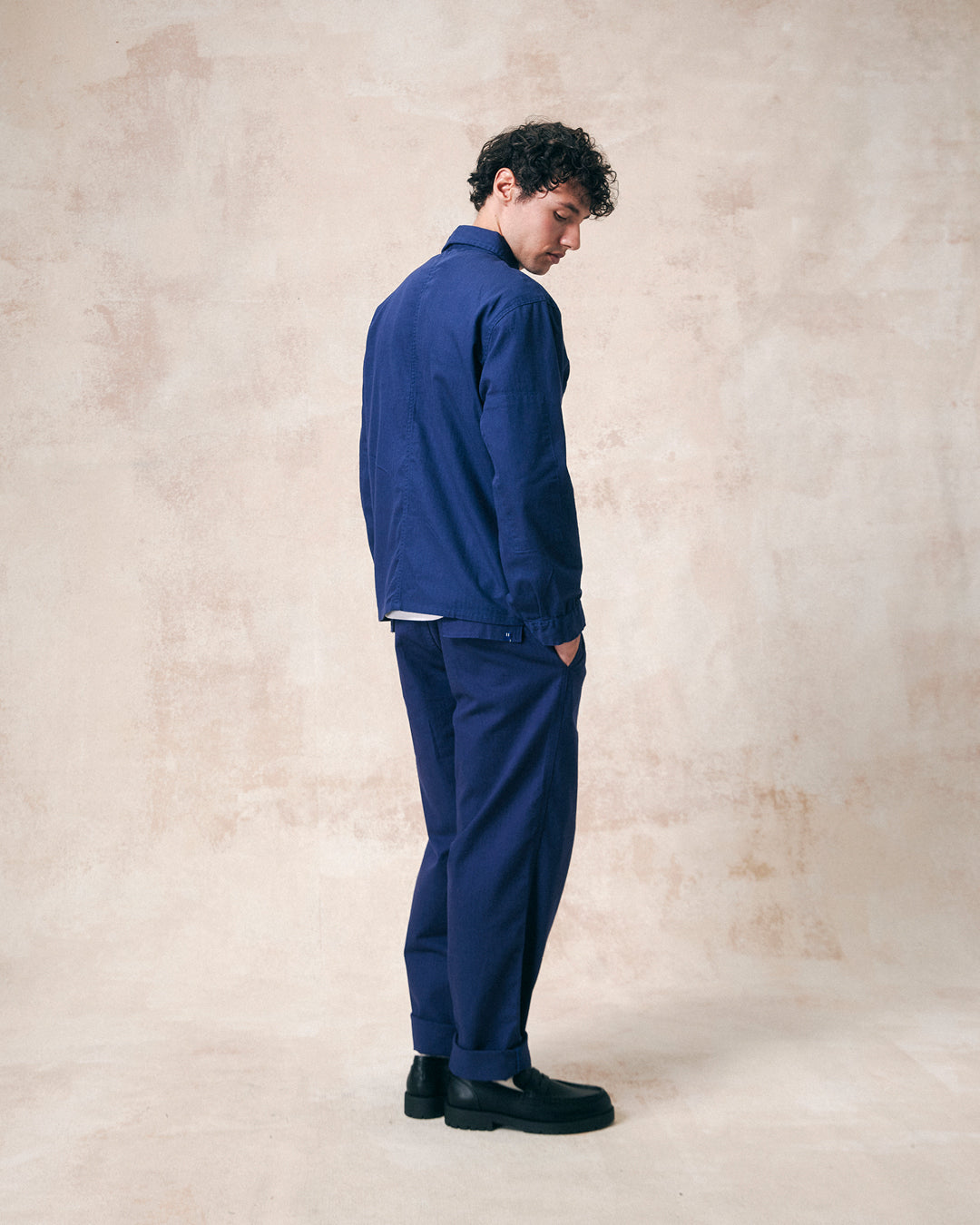 Full length model shot - angled back view of Uskees deep indigo organic herringbone cotton engineer jacket for men paired with herringbone barrel pants