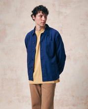 Front view of model wearing Uskees deep indigo organic herringbone cotton engineer jacket unbuttoned over a yellow t-shirt paired with khaki trousers.