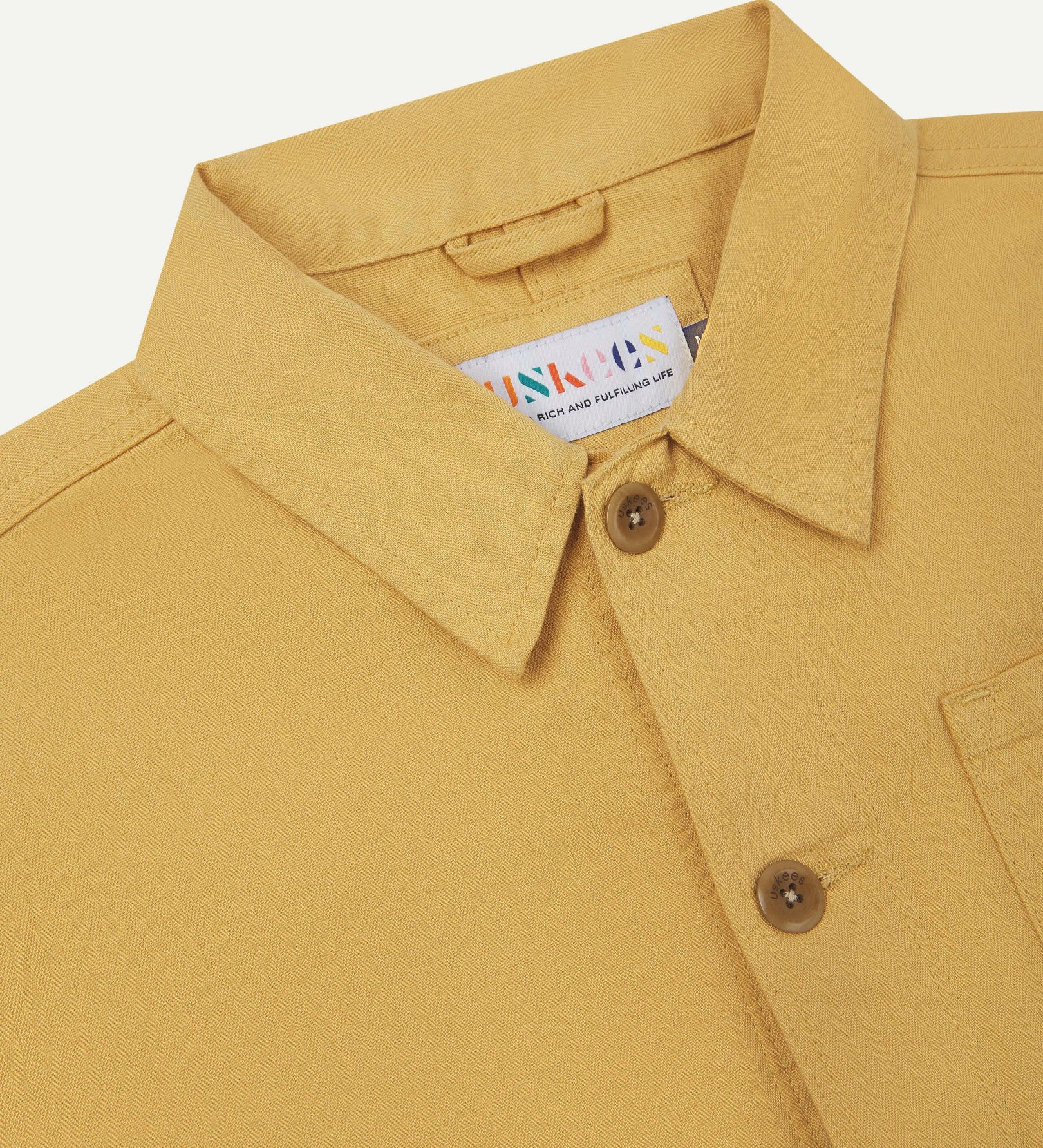 Neck view of Uskees 3040 sandstone-yellow herringbone cotton engineer jacket with focus on collar, Uskees brand label and matching corozo buttons.