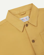 Neck view of Uskees 3040 sandstone-yellow herringbone cotton engineer jacket with focus on collar, Uskees brand label and matching corozo buttons.