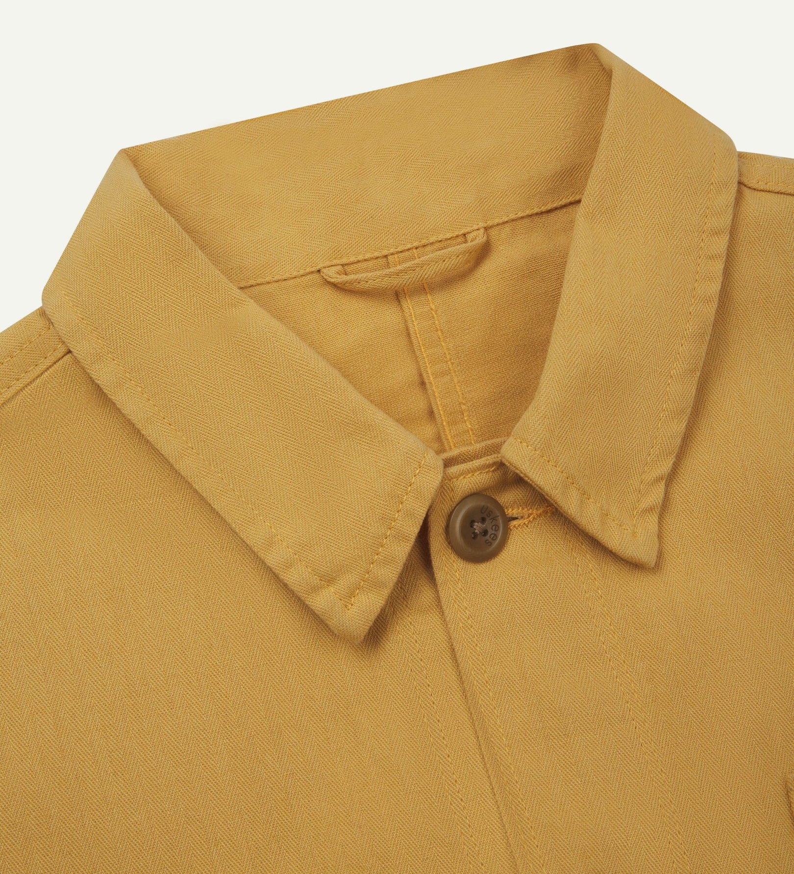 Neck view of Uskees 3040 sandstone-yellow herringbone cotton engineer jacket with focus on collar, Uskees brand label and matching corozo buttons.