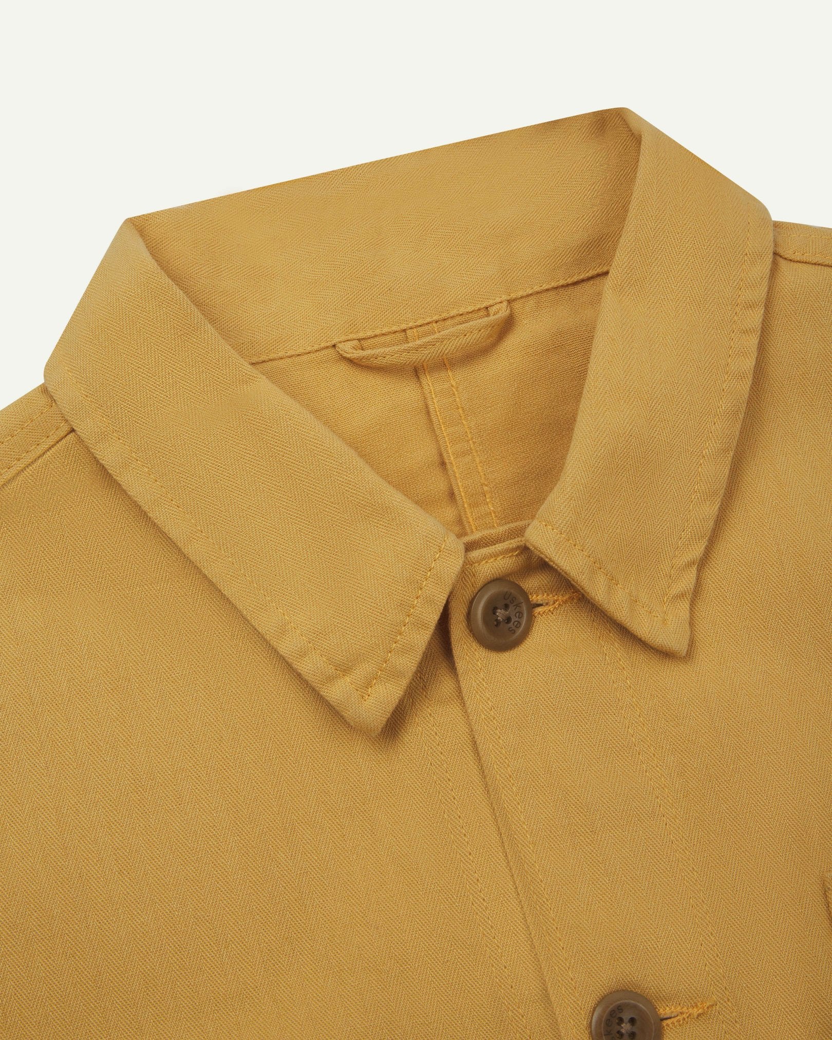 Neck view of Uskees 3040 sandstone-yellow herringbone cotton engineer jacket with focus on collar, Uskees brand label and matching corozo buttons.