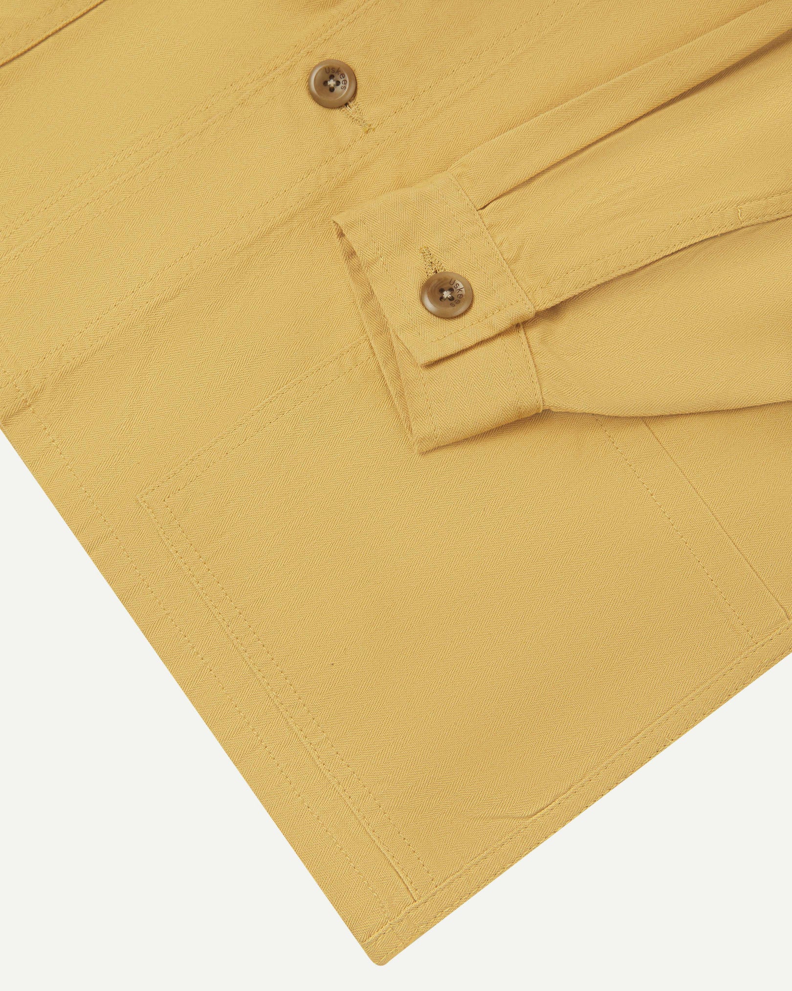 Close-up view of one of two hip pockets, cuff and Uskees branding tag of the 3041 signal jacket. Also is a closer view of the herringbone texture of the sandstone-yellow cotton.