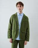 A male model wearing an Uskees coriander green utility-style blazer over a light blue button-down shirt, paired with matching  trousers, photographed against a plain light gray studio background.
