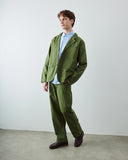 A male model wearing an Uskees coriander green suit consisting of a blazer and trousers, layered over a light blue dress shirt and paired with dark brown leather shoes, standing in a minimalist studio setting.