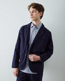 A male model wearing an Uskees navy blue blazer over a blue and red striped button-down shirt, standing against a light gray studio background.