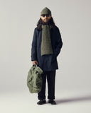 Male model wearing an Uskees midnight blue car coat, green scarf, and hat, holding a green bag against a plain background