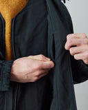 Close-up of a person wearing an Uskees charcoal lightweight shell jacket over a mustard yellow knit sweater, with both hands opening a side pocket.