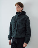 A male model wearing an Uskees dark charcoal hooded jacket with front flap pockets, standing against a plain light gray wall.