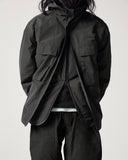 View of a male model from the neck down wearing an Uskees charcoal grey hooded waterproof shell jacket with the zip partially open from the bottom.