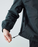 Close-up of a person wearing an Uskees dark charcoal gray lightweight jacket, pulling the elasticized drawstring hem down with their hand against a light gray studio background.