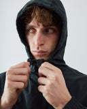 A close-up shot of a man wearing an Uskees charcoal hooded evo jacket, pulling the hood down slightly with both hands against a light gray studio background.
