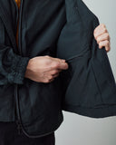 Close-up of a person wearing an Uskees charcoal lightweight jacket, opening the side panel to show a zippered pocket, with hands visible and a plain light gray background.