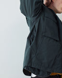Close-up side view of a person wearing an Uskees dark charcoal lightweight jacket, showing the open side pocket and shoulder area against a plain light gray background.