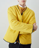 A man wearing an Uskees bright yellow liner jacket with black trim over a mustard yellow sweater and white collared shirt, photographed against a light gray studio background.