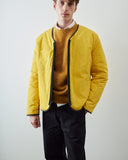 A man wearing an Uskees bright yellow zip front liner jacket with dark trim over a yellow sweater and white collared shirt, paired with black trousers, photographed against a plain light gray studio background.