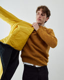 A man wearing a mustard yellow knit sweater and black trousers holds an Uskees bright yellow reversible liner jacket against his shoulder, photographed against a plain light gray studio background.