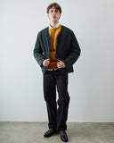 A man stands against a plain light gray wall wearing an Uskees dark charcoal quilted liner jacket over a mustard yellow knit sweater, a white collared shirt, black trousers, and black leather shoes.