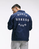 Top-half back view modelling Uskees 3052 navy blue coach jacket with focus on the the North Born Uskees slogan.
